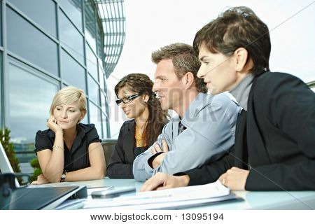 Picture or Photo of Group of young business people sitting in a row at table on office terrace outdoor, talking and working on laptop computer.